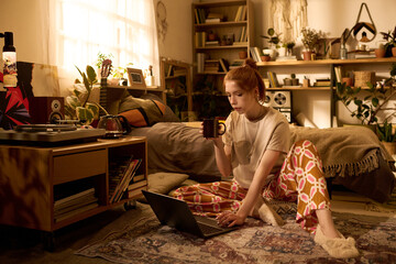Caucasian young adult woman sitting on floor using laptop and holding mug in cozy bedroom surrounded by plants and bookshelves, focused on screen while working or studying