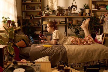 Young adult Caucasian woman lying on bed using laptop and holding bowl, surrounded by houseplants and bookshelves, studying or working in cozy bedroom with relaxed posture