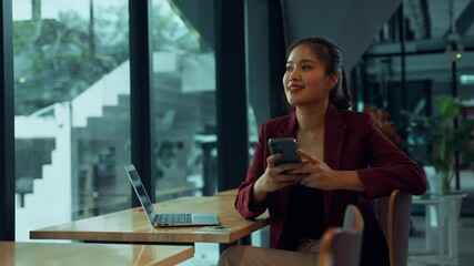Young successful asian businesswoman sitting at a cafe table with a laptop, happily texting on her smartphone and looking out the window - Powered by Adobe