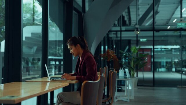 Focused asian businesswoman typing on her laptop keyboard, working in a modern office space or a contemporary cafe next to a window