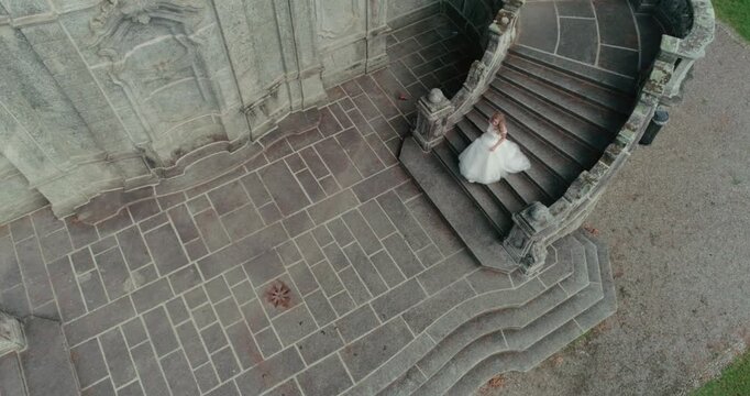 High-angle aerial view of a bride in a white wedding gown carefully descending a wide, ornate grey stone staircase of a historical building or park structure