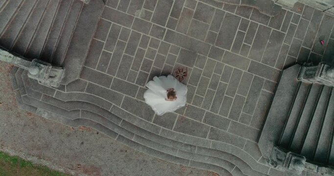 A high-angle drone shot looking down at a bride in a white wedding dress joyfully spinning on ancient, textured stone stairs. Captures movement, romance, architecture