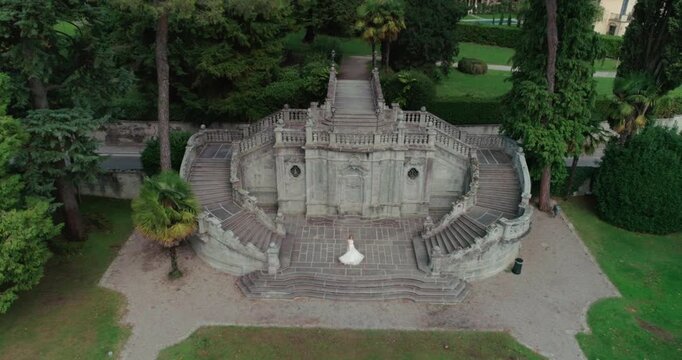 High-angle aerial view of a bride in a white wedding gown spinning at the base of a symmetrical, grand baroque stone staircase within a lush park or garden setting