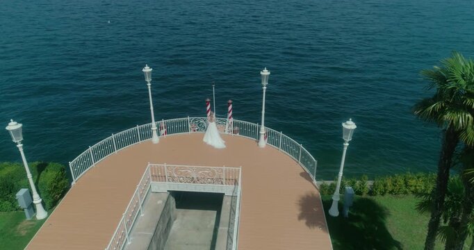 High-angle drone shot focusing on a bride in a white wedding dress standing on a decorated circular pier over deep blue lake water, with ornate white lampposts and palm trees framing the romantic