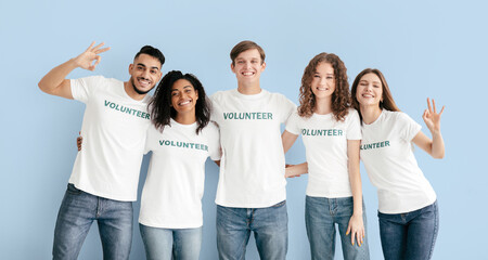 Five friends stand together against a light blue background, wearing matching white t-shirts with the word volunteer printed on them. They are smiling and making hand gestures.