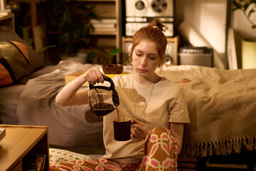 Young adult Caucasian woman sitting on floor pouring coffee from glass pot into mug in cozy bedroom setting, focused expression, relaxed posture, casual home environment