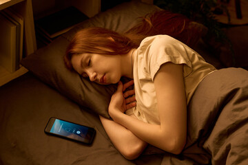 Caucasian young adult woman sleeping on bed with smartphone displaying incoming call screen beside her on pillow, resting with eyes closed and hand near face in dimly lit bedroom