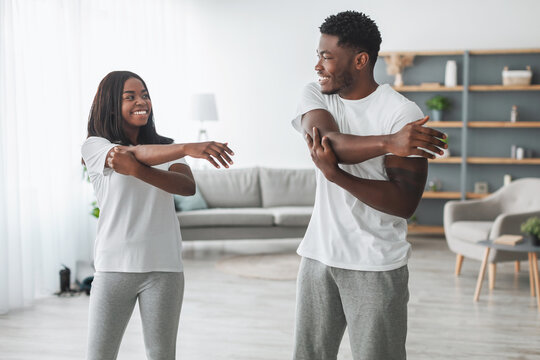 Warm Up Concept. Portrait of happy young African American couple stretching arms before home training, standing in living room and looking at each other, smiling guy and lady exercising together - Powered by Adobe