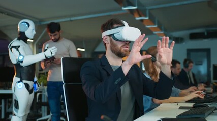 Male operating object in virtual reality headset. Coworkers focusing on tasks at computers nearby. Humanoid robot interacting with another male holding tablet. Active office environment. - Powered by Adobe