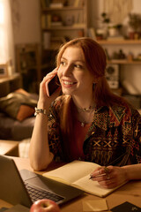 Caucasian young adult woman smiling while talking on smartphone and writing in notebook, sitting at desk with open laptop in home office setting, looking away from camera
