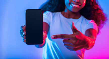 A smiling woman holds a smartphone in front of her while pointing to the screen. The background is illuminated with vibrant blue and pink lights, creating a playful atmosphere.