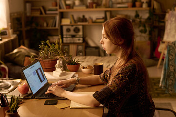 Caucasian young adult woman working on laptop at desk in home office, focusing on screen while typing, surrounded by plants and bookshelves, studying or remote working