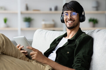 A young man with glasses and headphones is sitting comfortably on a sofa, using a tablet. He appears happy and focused on the content, enjoying a leisurely moment in his home.