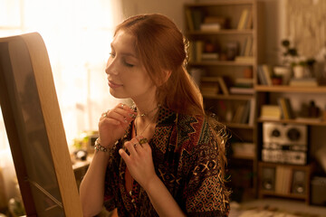 Caucasian young adult woman adjusting necklace while sitting at vanity mirror, long red hair styled back, serene expression, surrounded by bookshelves and personal items