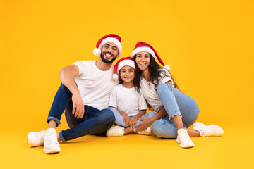 A joyful family sits together on a vibrant yellow backdrop, all wearing Santa hats. They share...