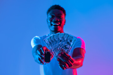 A person stands against a colorful background, joyfully displaying a handful of cash in one hand...