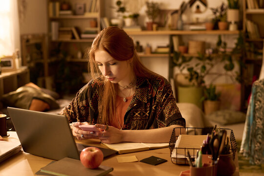 Young adult Caucasian woman sitting at desk using smartphone while working on laptop, surrounded by stationery and plants, focusing on multitasking in home office environment - Powered by Adobe