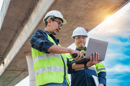 Construction Workers Using Laptop for Project Management Under Bridge with Sunlight and Cloudy Sky