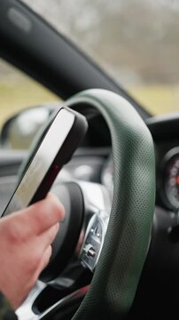 The driver, while driving a car, uses a mobile phone, types text in a chat, and violates traffic rules. Closeup Steering Wheel Hand On Rim, Driver Adjusting Grip And Focusing On Road and phone