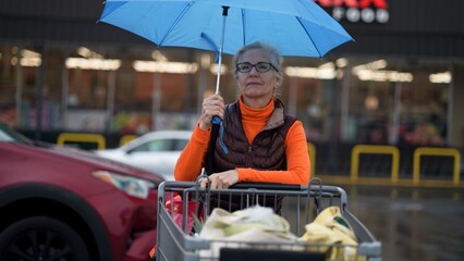 Mature woman looks at different grocery items with an umbrella in her hand outside a store on a rainy day.