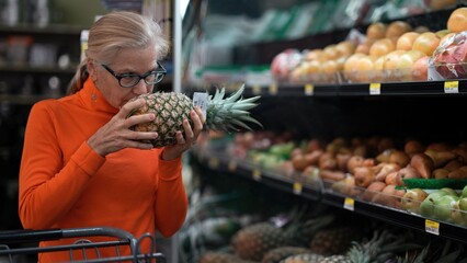 A mature woman inspects a pineapple while comparing it with other fruits in a grocery store. She is focused on making a choice.