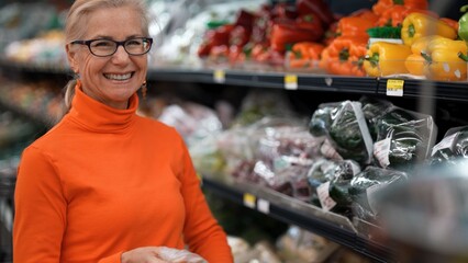 Mature woman smiles while comparing prices of fresh produce in a supermarket. She looks at different food options for her shopping list.