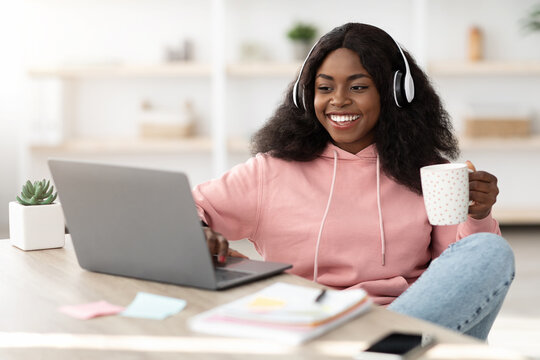 A cheerful young woman wearing a pink hoodie enjoys her study session at home. She listens to music with headphones on and holds a cup while typing on her laptop. - Powered by Adobe