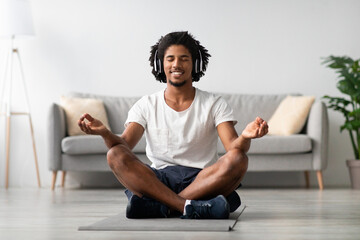 A relaxed African American man sits in a lotus position on a yoga mat in his living room, wearing wireless headphones and meditating with closed eyes.