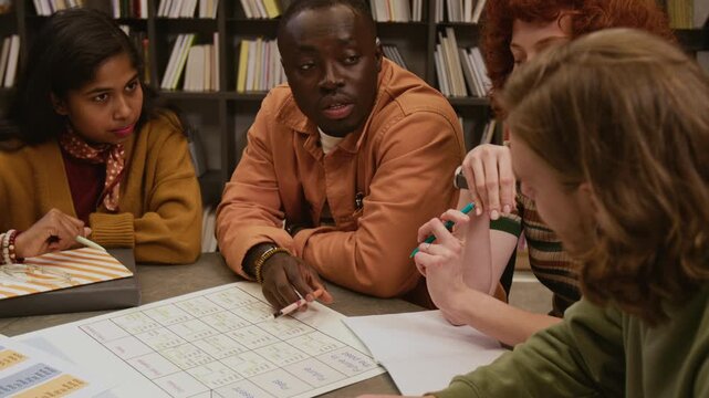Tilt up shot of African American male teacher pointing at irregular verb chart explaining English grammar rule to group of adult students gathered at library table in language course for beginners