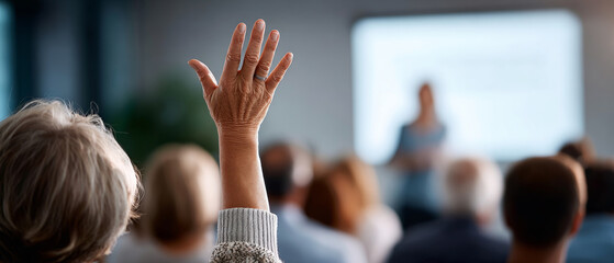 Senior person raising hand to ask question during a seminar or workshop with blurred presenter and audience in background