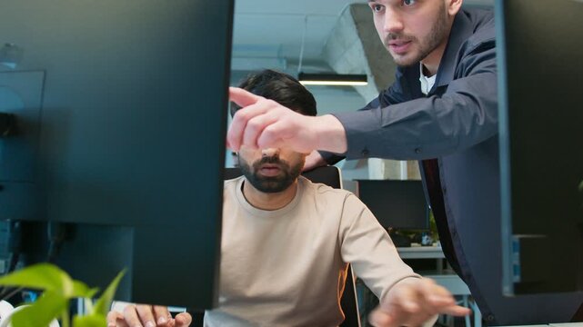 Coworker pointing out mistake in code on monitor. Male fixing issue while listening. Two teammates solving problem together during development task. Office environment supporting focused workflow.