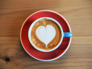 A top-down view of a blue coffee cup with a heart-shaped latte art on a red saucer, placed on a wooden table.