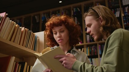 Low angle shot of focused young woman reading through pages of textbook with male groupmate while choosing educational material in library aisle
