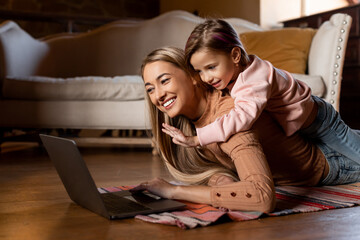 A mother and her young daughter are having fun together. The girl is playfully resting on her mother's back while they explore a laptop on the floor of their warm living room.