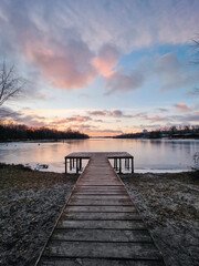 Wooden pier on the lake (wide angle diminishing perspective view). Winter sunrise scene with ice over the water, trees and sky reflecting in mirror-like lake surface.