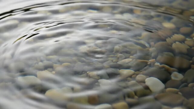 Close-up of a water drop creating ripples on a pebble-filled surface.