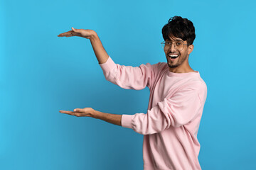 A cheerful young man stands in front of a bright blue background, smiling widely while gesturing with both hands as if presenting something exciting.