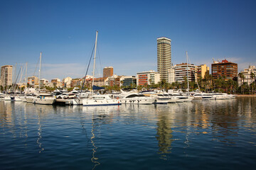 Waterfront lined with palm trees and both historic and modern buildings along the Explanada de Espana and marina with yachts reflecting in the mediteranean sea. City of Alicante, Costa Blanca, Spain.