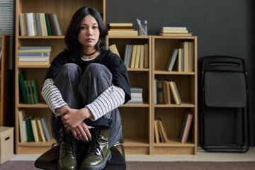 Portrait of young Asian woman sitting on chair with knees drawn to chest looking at camera during group therapy session with bookshelves in background © Mediaphotos