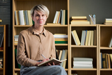 Portrait of adult woman sitting with notebook and pen, looking into camera, participating in group therapy session, bookshelves in background, short blond hair