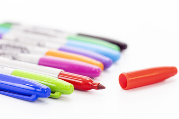 Close-up of colorful markers lined up on a white background with an uncapped red marker and its red cap beside it