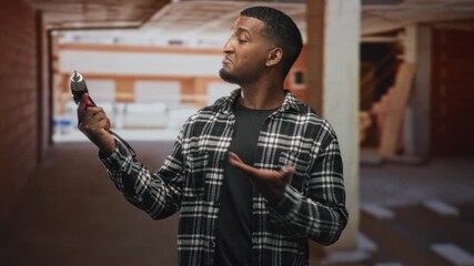 Man holding electric screwdriver and gesturing with hand in building interior, wearing plaid flannel shirt while inspecting the tool; confidence skill.