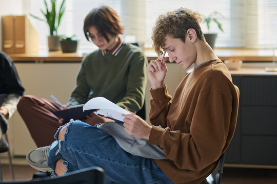 Teenage boy sitting in group therapy session reading document while another teenage boy with dark hair writing in notebook in background, both focused on activity