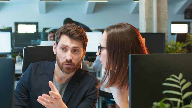 Caucasian male sitting at desk asking questions while gesturing toward monitor. Female supervisor listening closely while reviewing code. Modern and bright office workspace. Giving instructions.