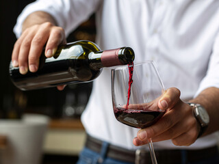 A person's hands pouring red wine from a dark bottle into a clear wine glass.