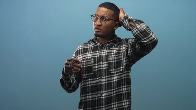 Man holding car keys and remote fob, wearing glasses and plaid shirt, hand to head with puzzled expression in studio; uncertainty thought.