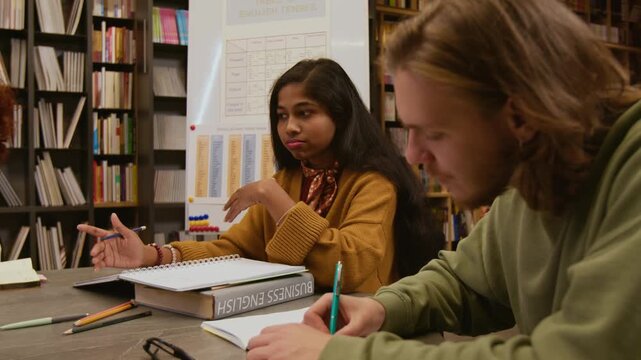Side view of adult male student writing in notebook while young Indian female teacher explaining irregular verbs to group of learners during library based business English session