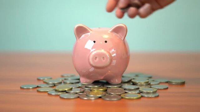 A pristine pink ceramic piggy bank receives sparkling silver coins dropped by a caucasian hand on polished wood, soft blue background. Concept of consistent financial saving