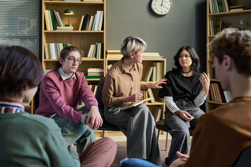Diverse group of young people and teenagers sitting in circle in group therapy session, adult woman facilitating discussion, bookshelves in background © Mediaphotos