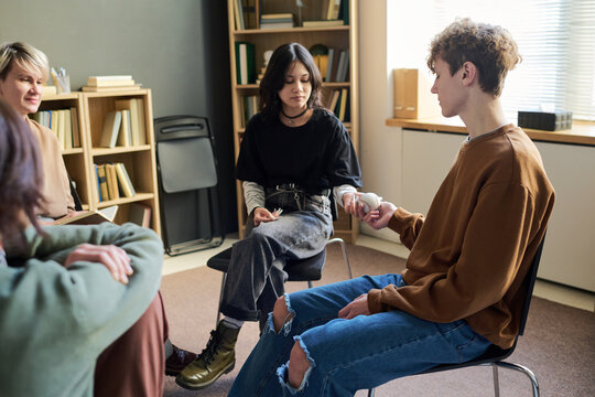 Diverse group of teenagers sitting in circle participating in group therapy session, teenage boy handing speaking ball as token to Asian teenage girl, others attentively observing interaction - Powered by Adobe
